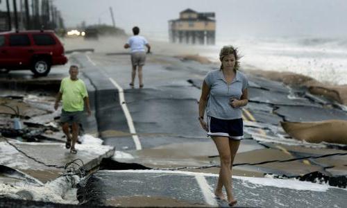 Three people walk on a road that's been broken apart by a storm. 