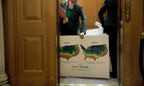 A woman entering a hearing holding a sign that shows two maps that compare plant hardiness zones.